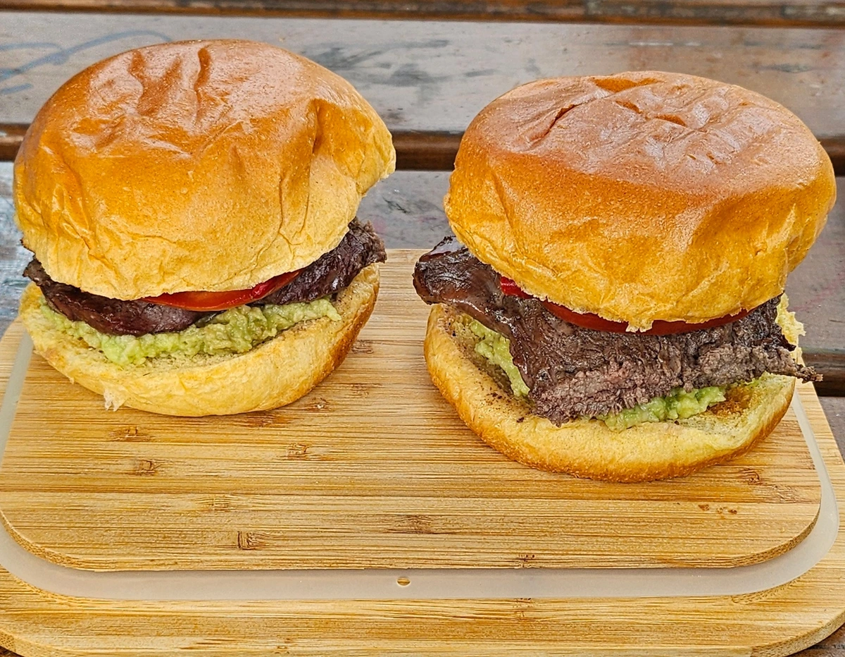 Two churrasco sandwiches on a wooden board, containing avocado paste, tomato, mayonnaise, chili and grilled beef