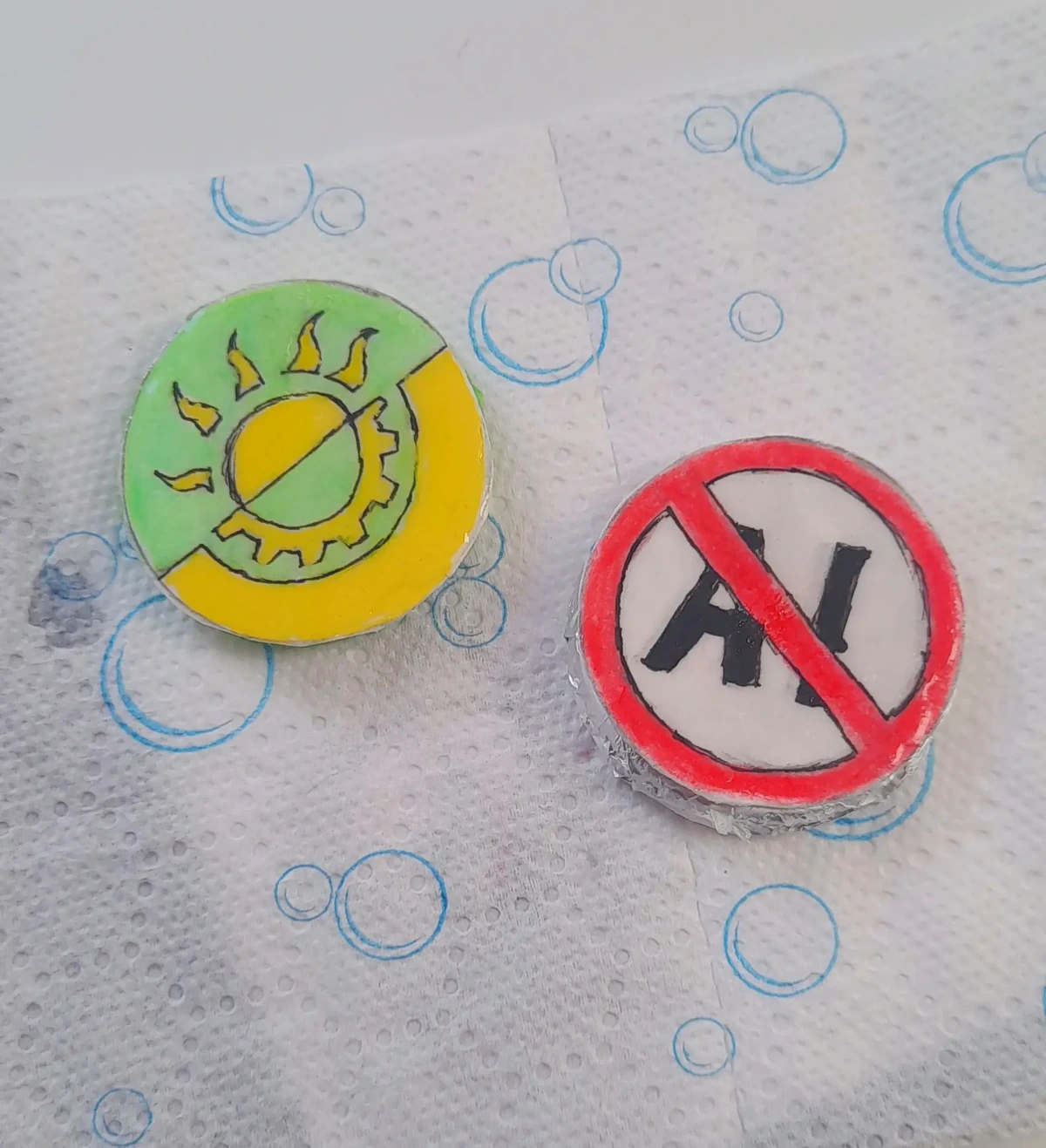 Two pins I made, one is showing a solarpunk symbol (yellow and green flag separated diagonally, like anarchist flags, with a half of a sun symbol, representing solar power, on the green side and a half of a gear symbol on the yellow side, representing technology) and the other one has "AI" written on it, crossed out with a red stroke